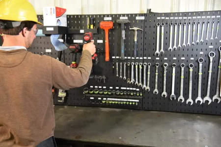 A person in a yellow hard hat and safety glasses uses a power drill in front of a wall-mounted tool board, demonstrating the 5 Key Features of a 5S Workstation with neatly organized wrenches, hammers, and other hand tools.