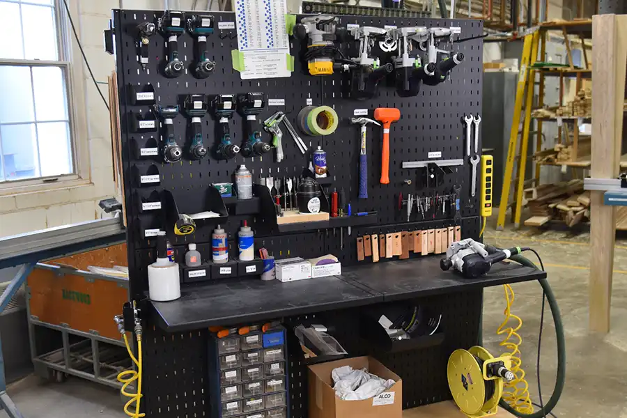 A workshop pegboard displays drills, hammers, tape, and clamps—plus shelves with bottles and drawers—showcasing the 5 key features of a 5S workstation in an organized, well-lit space with boxes of parts on a black workbench.