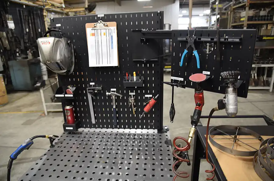 A metal workbench with a perforated tool board displays various tools—pliers, screwdrivers, a welding helmet, and more—demonstrating the 5 key features of a 5S workstation in an organized industrial workshop setting.