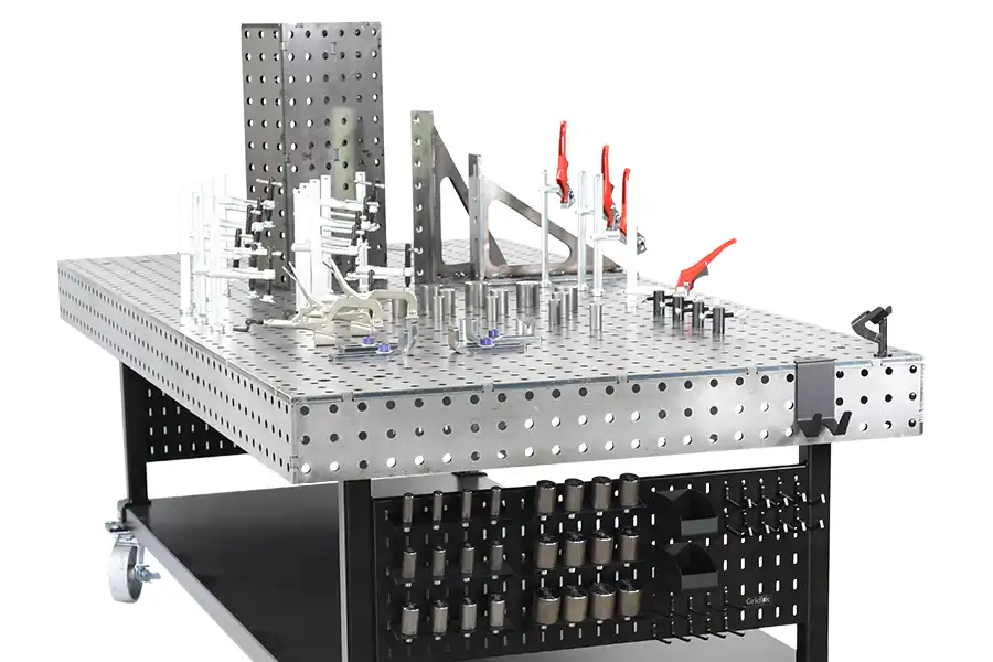 A metal welding table with a grid of holes, various clamps, brackets, and fixtures attached, and tools stored on a lower shelf and rack. The table is on wheels for mobility.