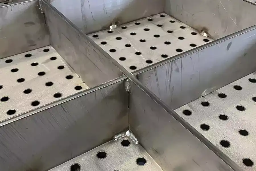 Close-up of metal trays with perforated bottoms and high edges, divided into sections. The metal surfaces show some scratches and spot welds at the corners—signs often seen on a well-used welding table.