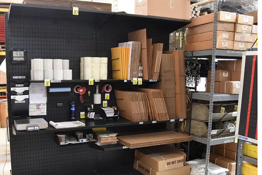 A warehouse packing station showcases 5 key features of a 5S workstation, with shelves holding shipping labels, tape, scissors, boxes, mailing tubes, and other supplies—all neatly organized and clearly labeled for efficiency.